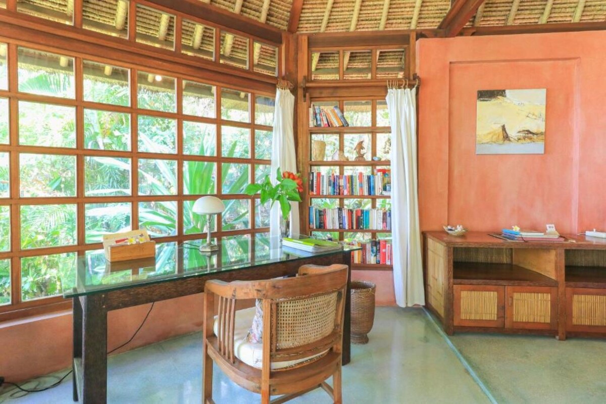 A room with a glass table, wooden chairs, a bookshelf, and a window with a view of greenery.