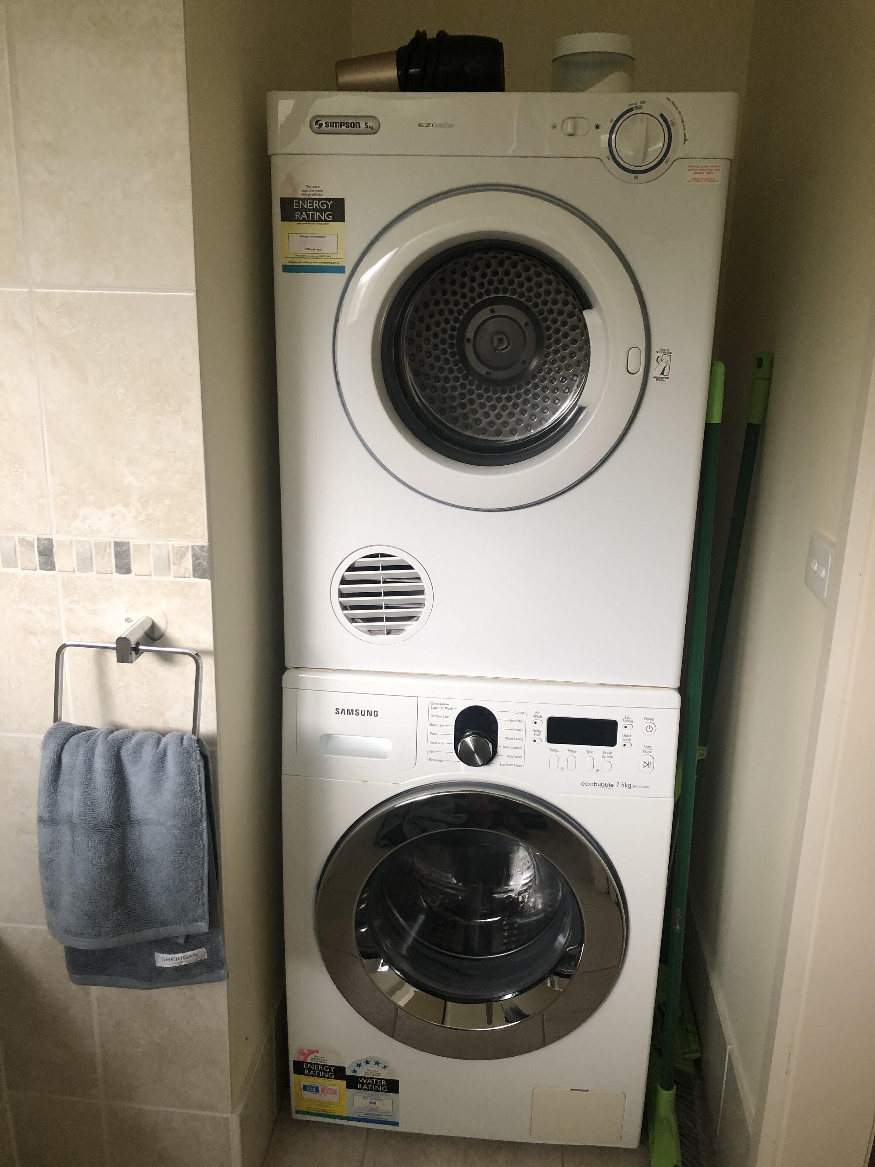 A stacked washer and dryer unit with energy and water rating labels, a control panel, and a towel rack with a blue towel.