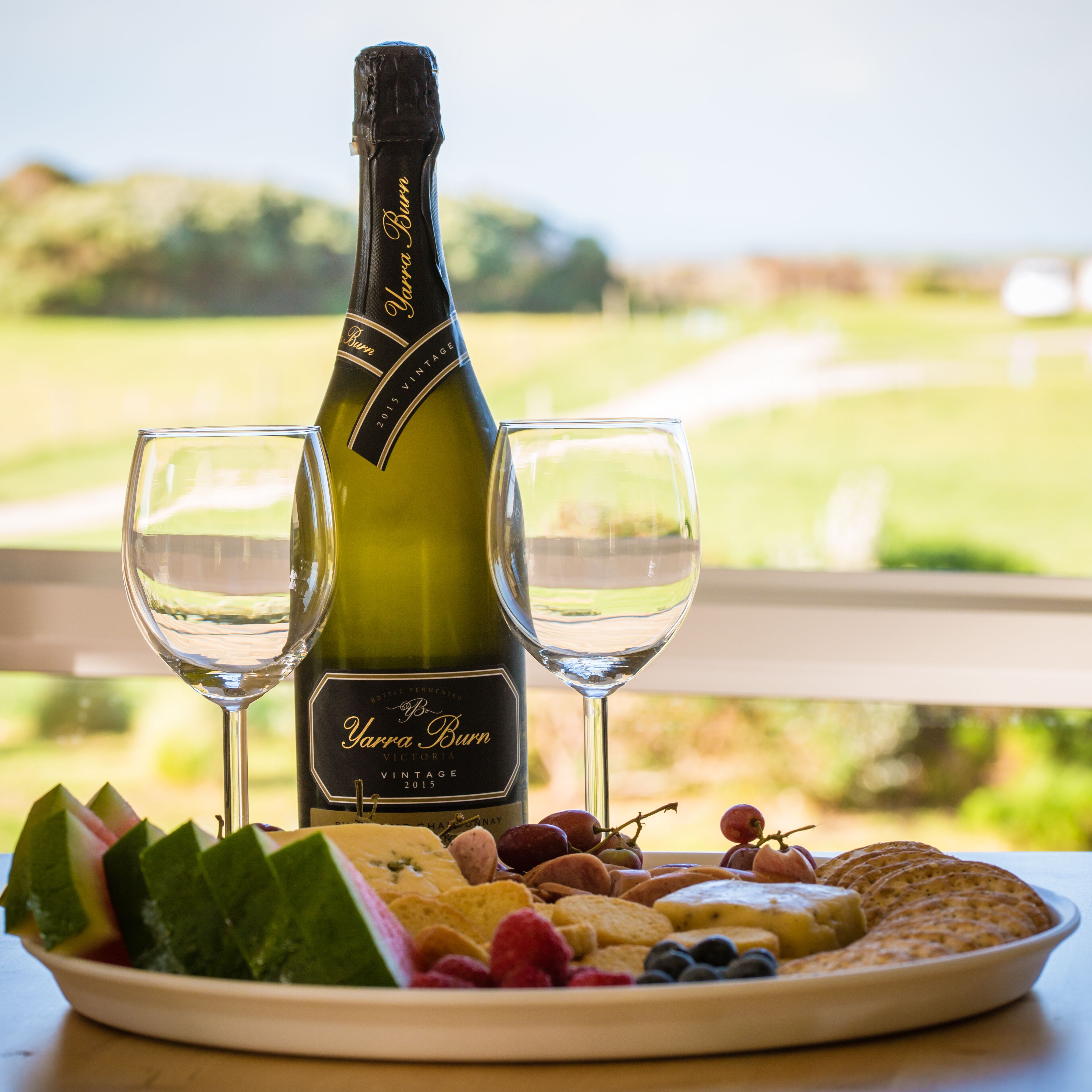 A bottle of Yarra Burn 2015 vintage wine, two glasses, and a fruit platter on a table with a view of a golf course.