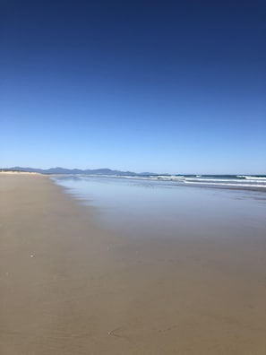 Une vaste plage de sable fin, avec de douces vagues et des montagnes lointaines sous un ciel bleu dégagé.