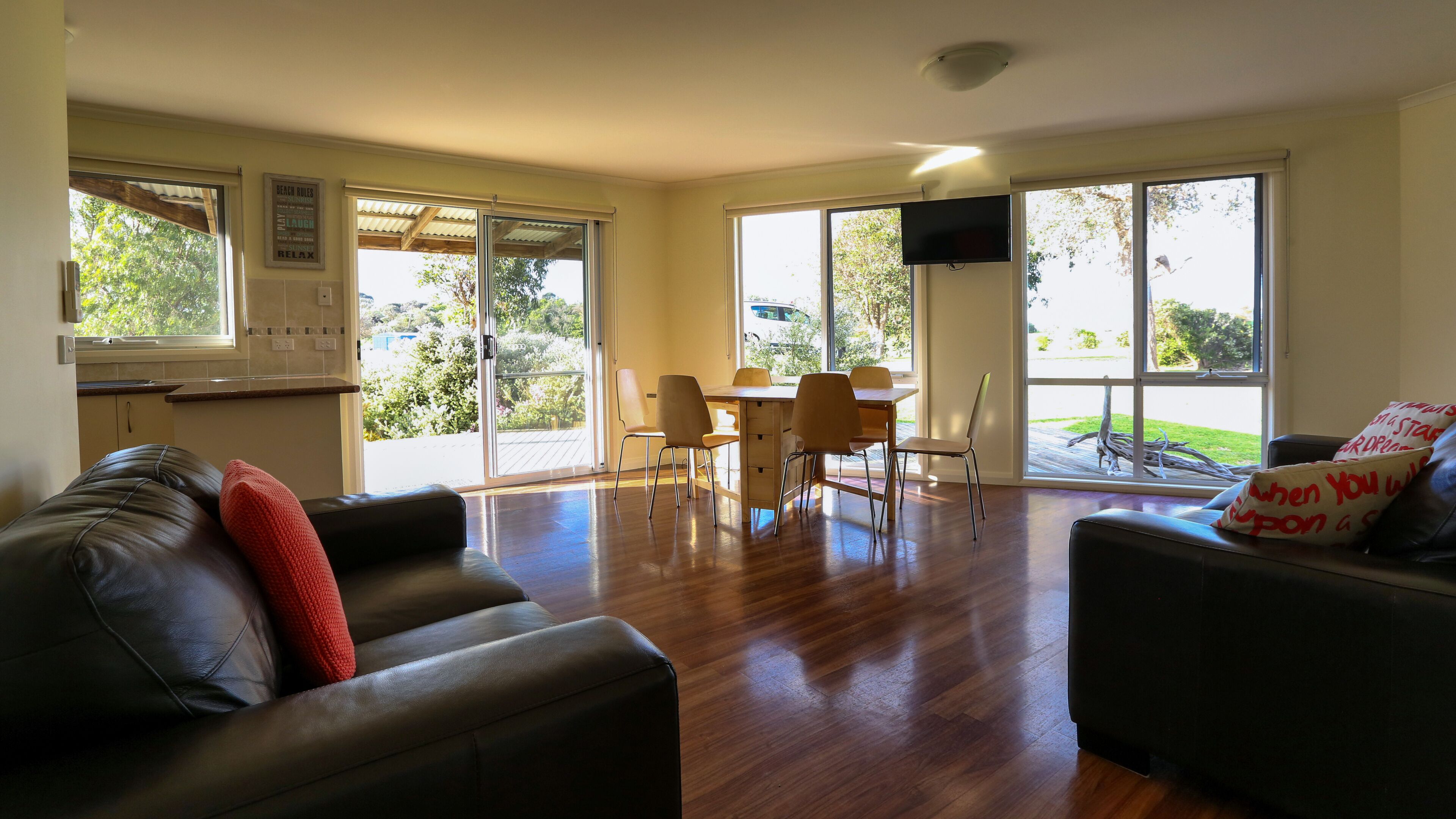 A spacious living area with a wooden floor, a black leather sofa, a dining table with chairs, and large windows offering a view of the outdoors.