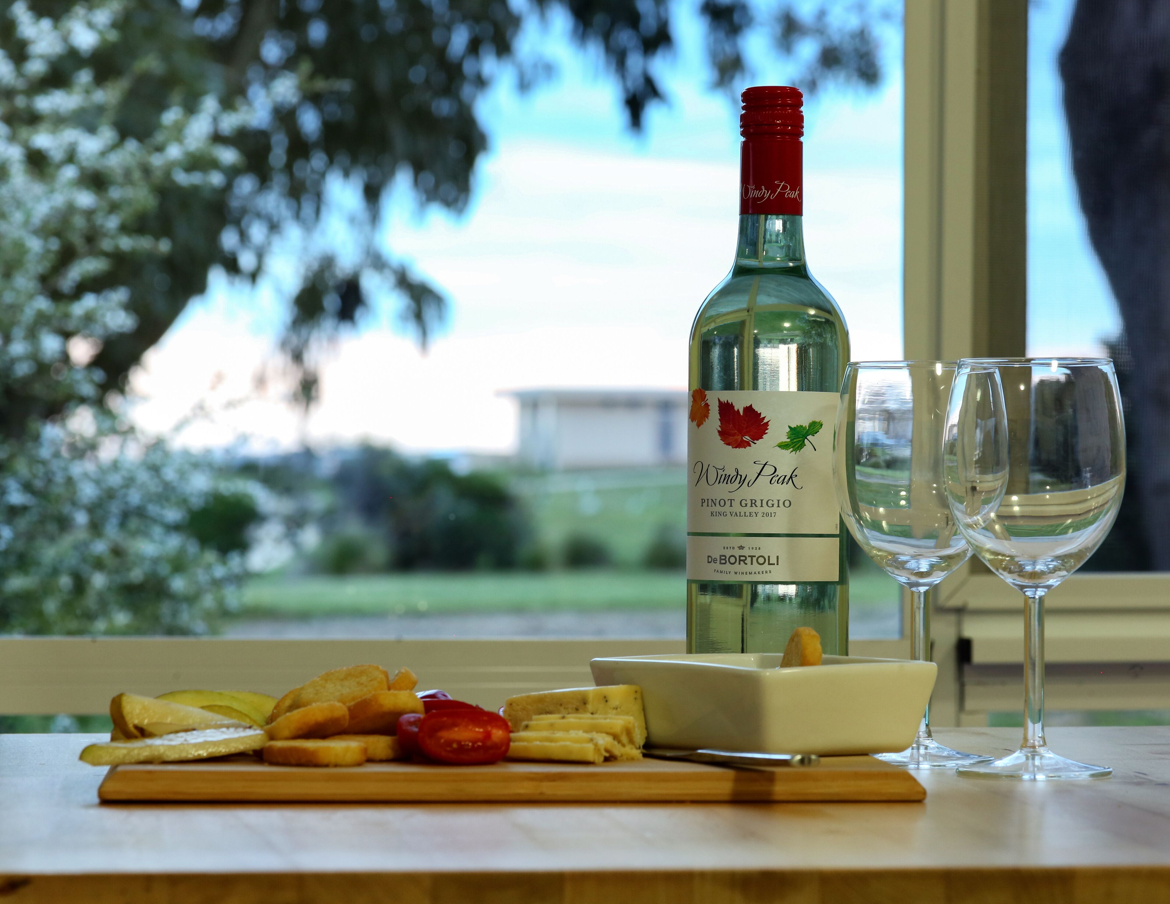 Una ampolla de vi Pinot Grigio Windy Peak King Valley 2017, dues copes de vi buides i una taula de fusta amb pa, tomàquets i formatge.