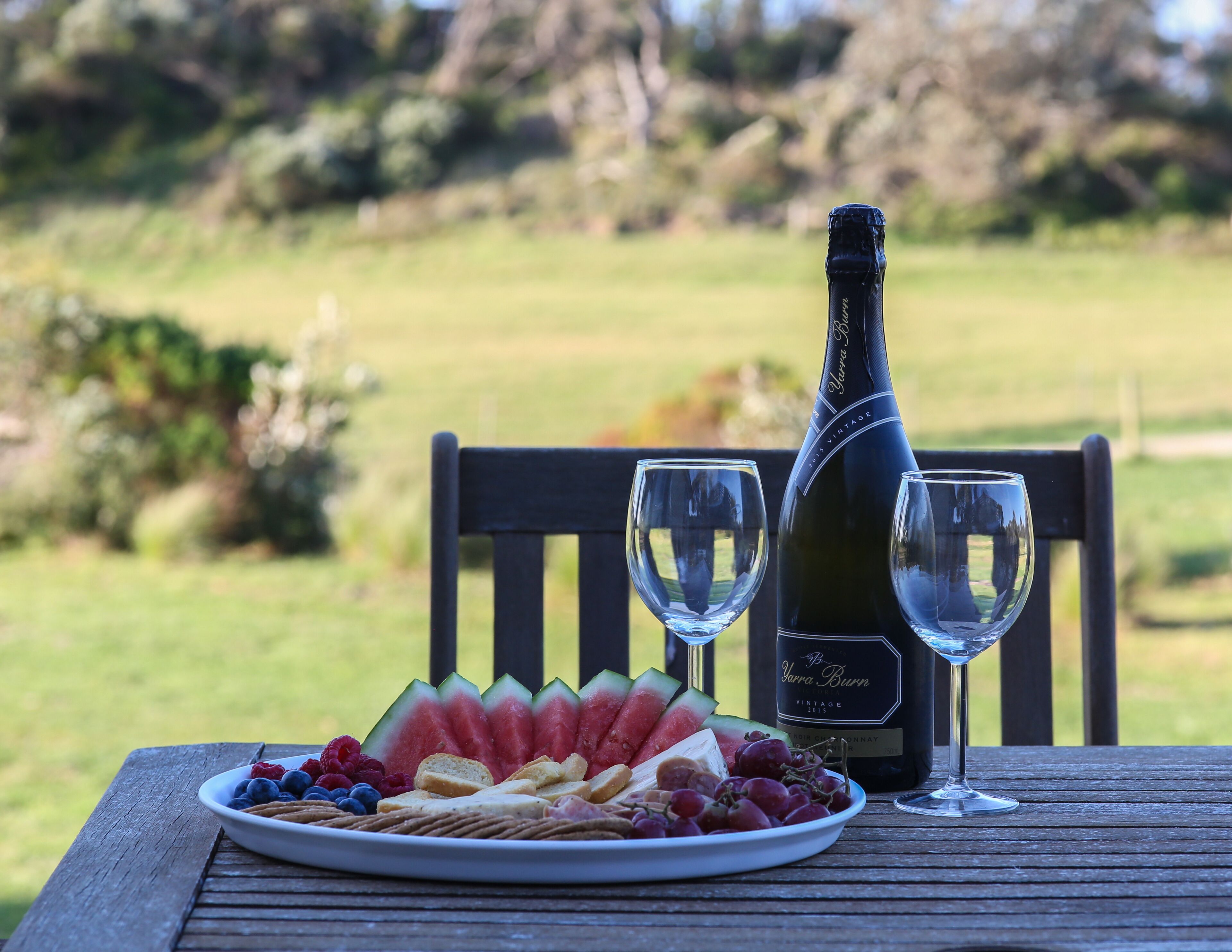 Una botella de vino, dos copas y un plato de comida sobre una mesa de madera al aire libre.