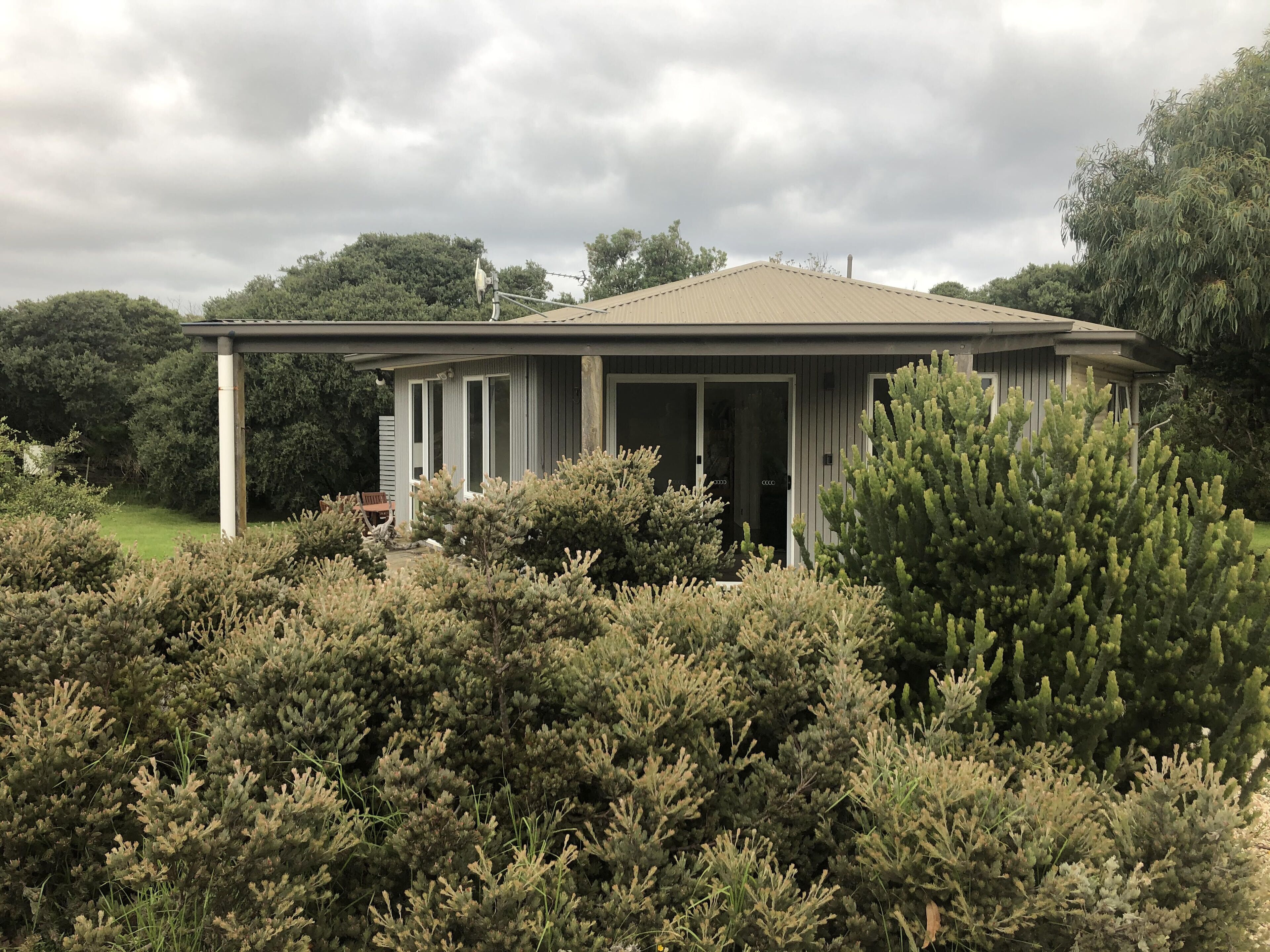 Een gelijkvloers huis met een overdekte veranda, omgeven door weelderige struiken en bomen.