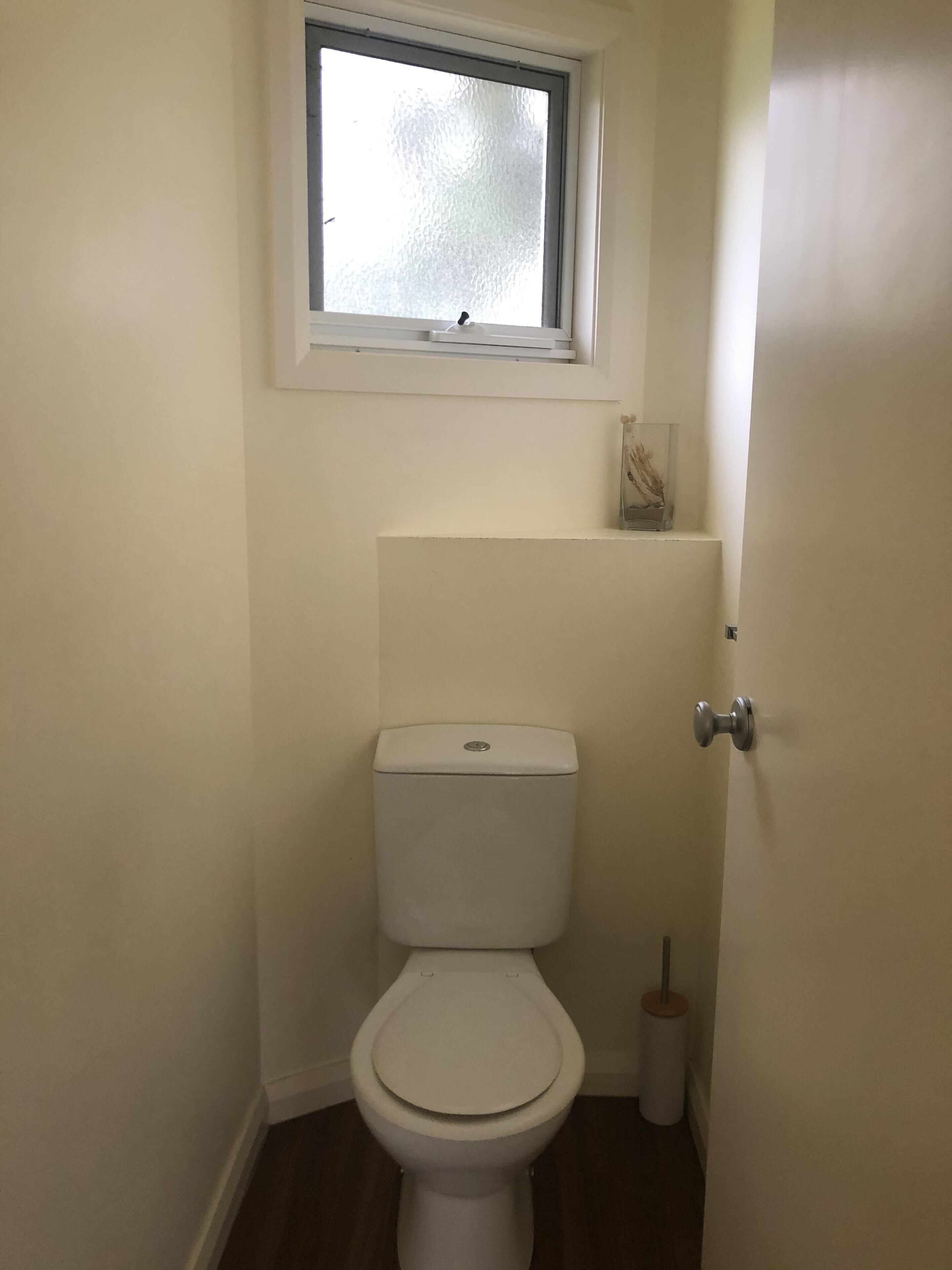 A small, white toilet with a window above it, a small shelf with a decorative item, and a toilet brush.