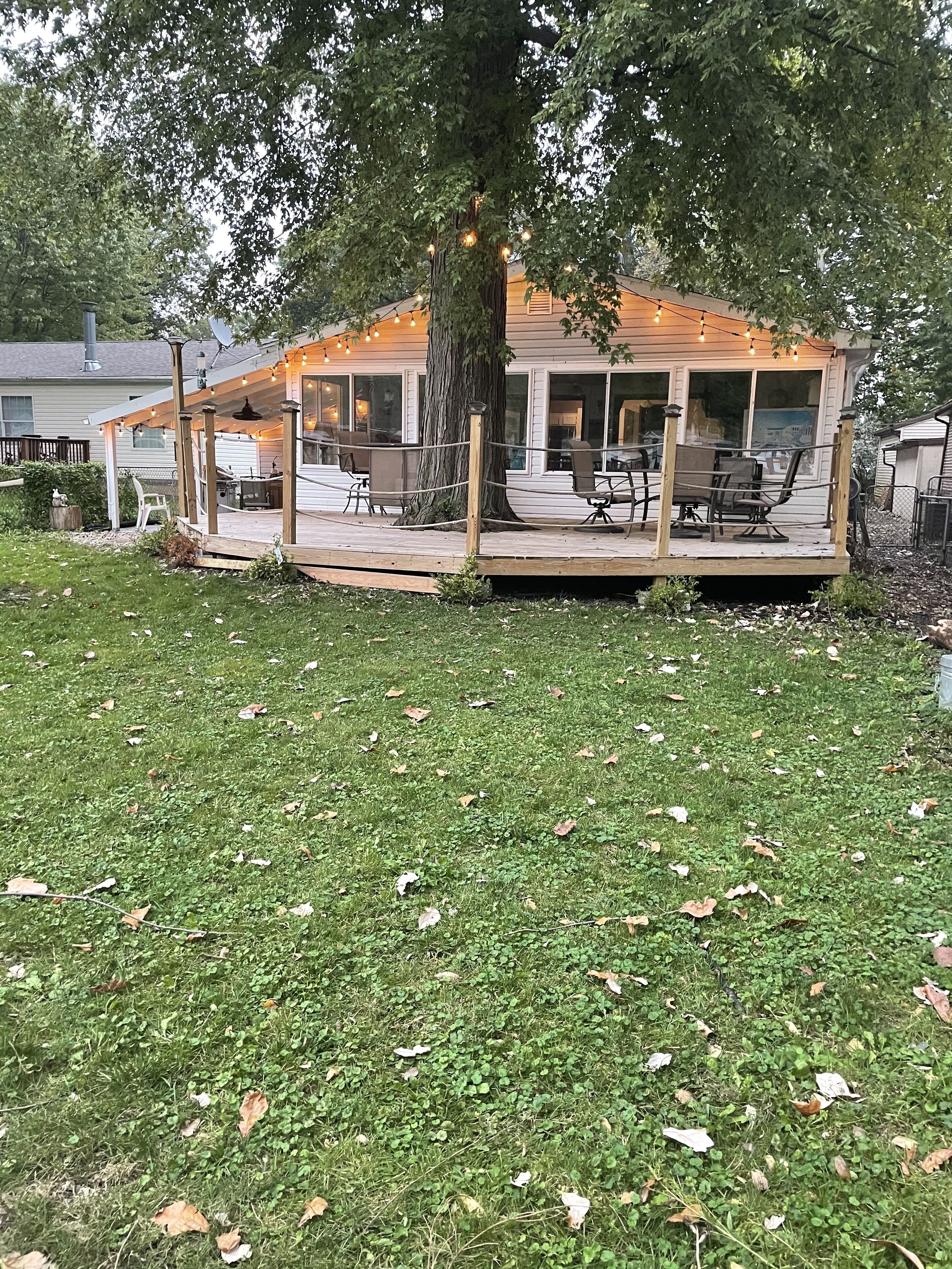 En overdækket terrasse med siddepladser og lyskæder, omgivet af græsplæne og træer.