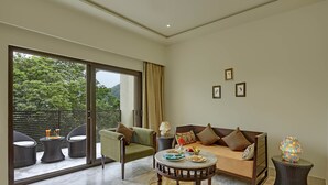 A modern living room with a sofa, armchairs, a coffee table, and a dining table. There are two lamps, a window with curtains, and a view of greenery outside. - The Kumbha Residency-Luxury Resort and Spa (Kumbhalgarh)