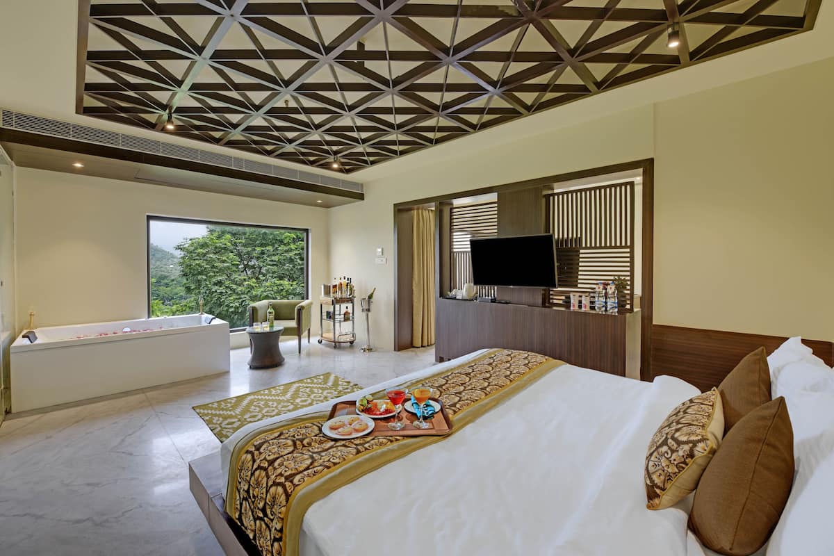 A hotel room with a large bed, a bathtub, a TV, and a view of greenery.