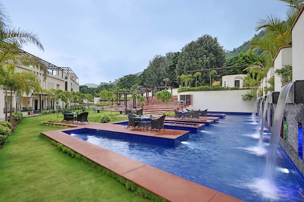 Zona de piscina con sillones y mesas, rodeada de vegetación y un telón de fondo montañoso.