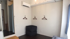 A minimalist room with a dark wooden floor, a black ottoman, and a bed with grey bedding. There are three wooden hangers on the wall and an air conditioning unit above. - GOLDEN MILE Hostel (Amami)
