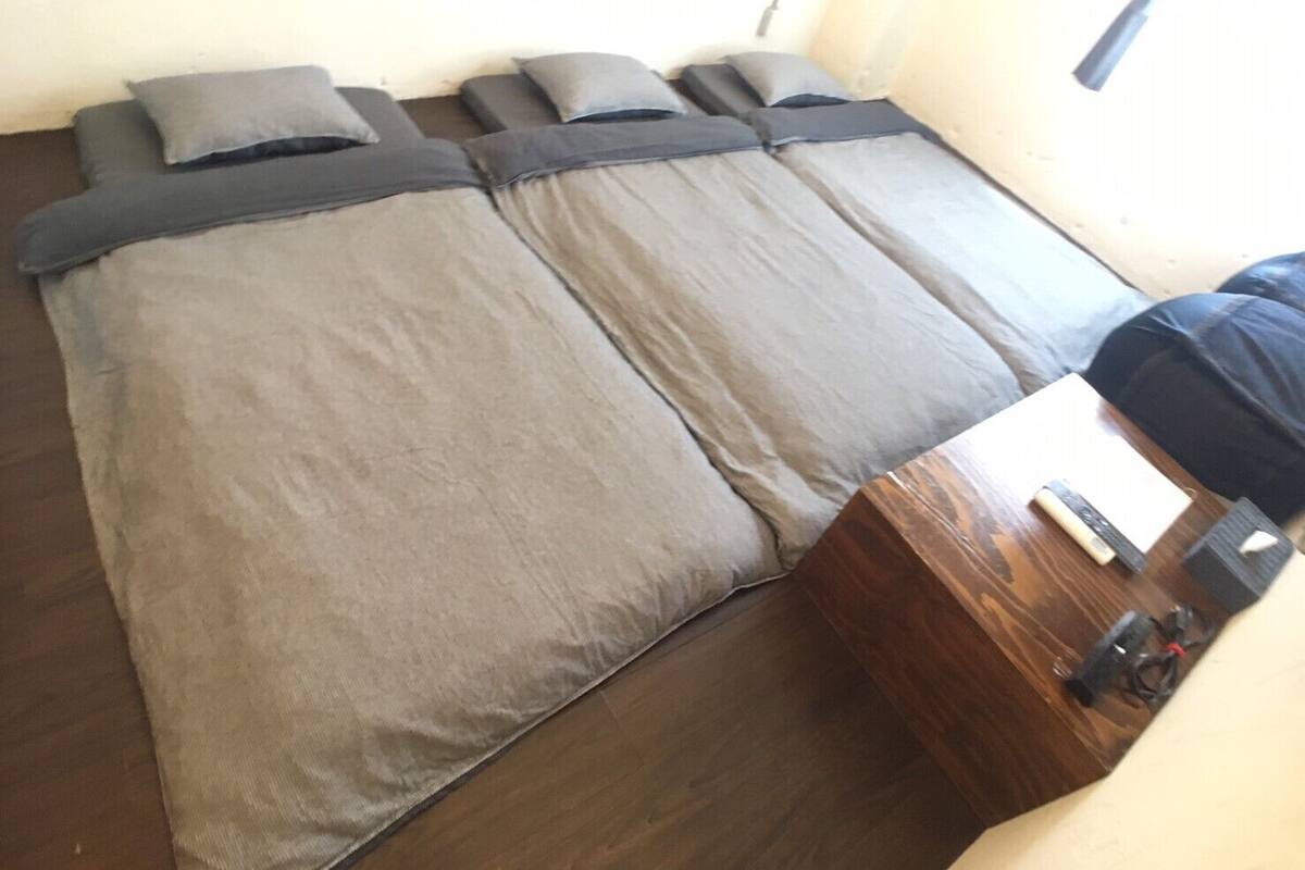 Une chambre avec un grand lit recouvert d’une literie grise, une table de chevet en bois et une fenêtre équipée de stores.