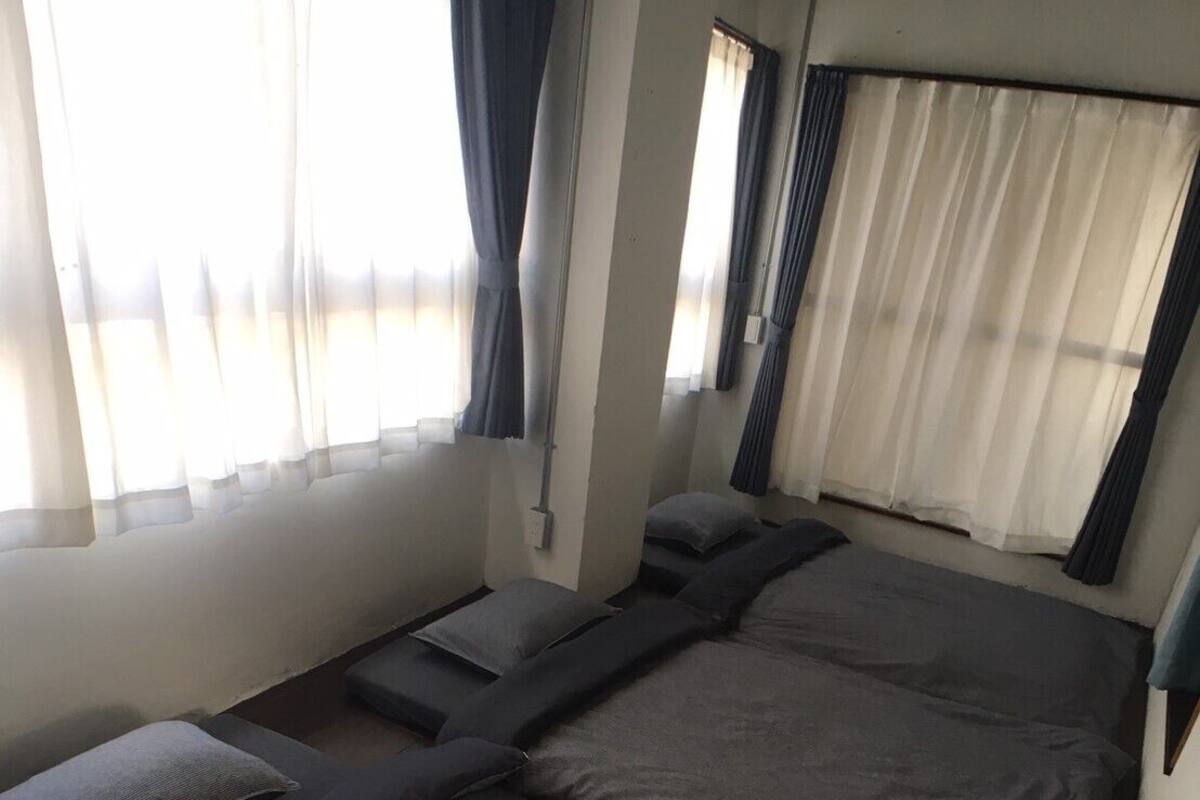 Une chambre avec deux lits, chacun étant équipé de draps et d’oreillers gris. Il y a des fenêtres avec des rideaux légers et des rideaux plus épais.
