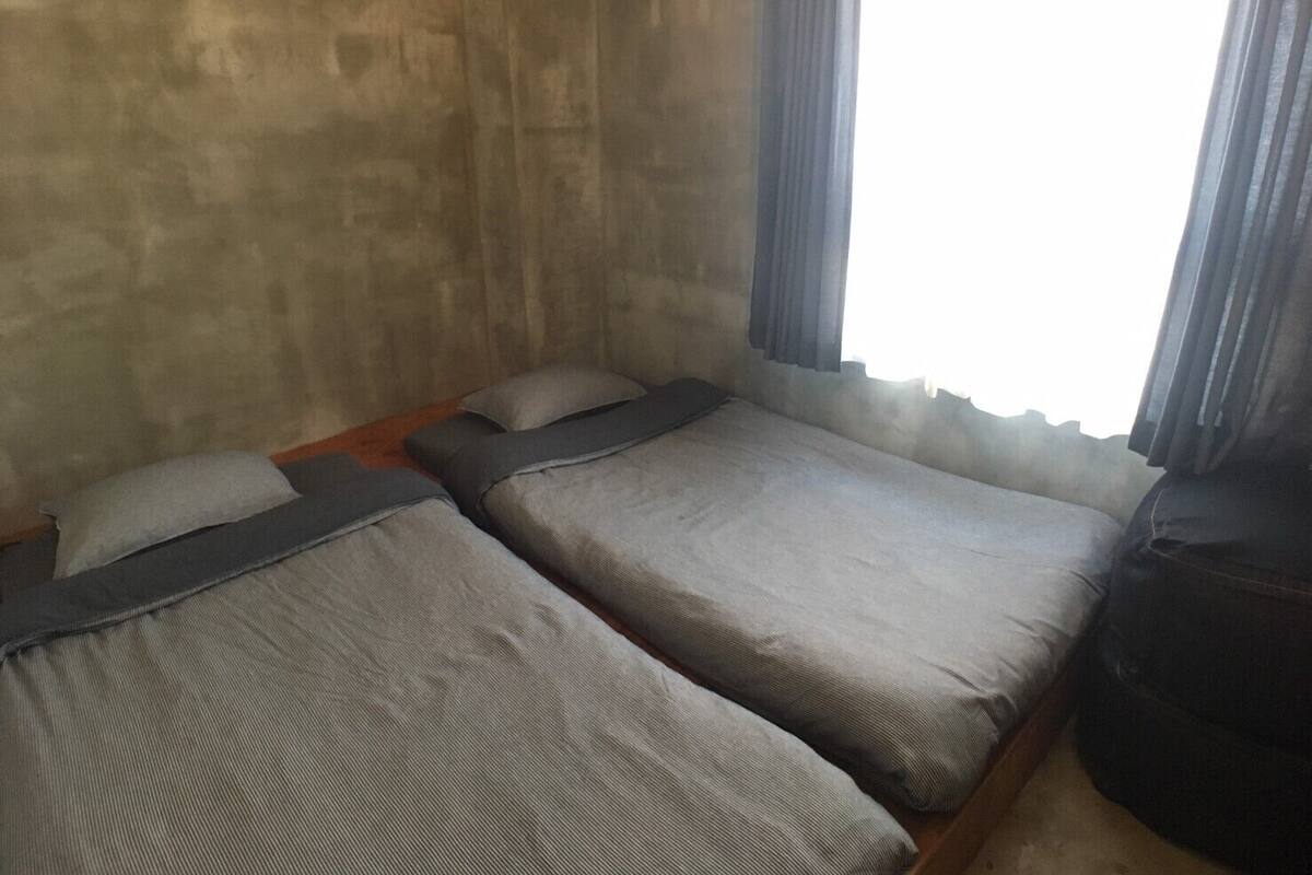 Un petit logement composé d’une seule pièce, avec deux lits, un plancher en bois et une fenêtre avec des rideaux.