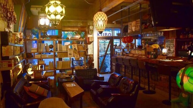 A cozy, dimly lit bar area with wooden shelves, leather seating, and hanging lamps. - GOLDEN MILE Hostel (Amami)