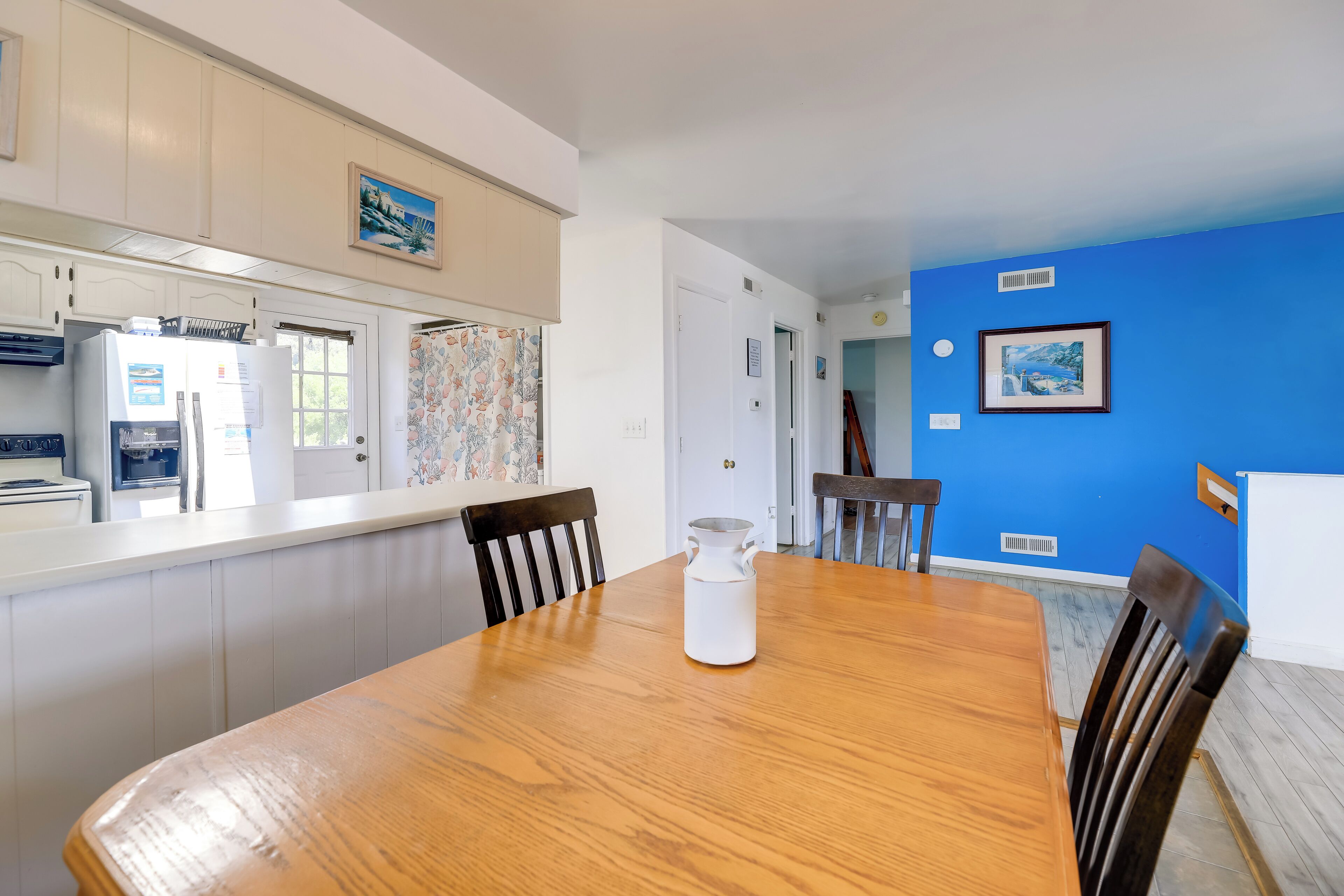 Un comedor con una mesa de madera, sillas y una jarra blanca. Se observa una cocina con gabinetes blancos y un refrigerador. Las paredes están pintadas de azul y blanco, y hay cuadros enmarcados.