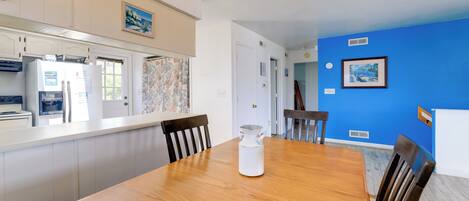 Un comedor con una mesa de madera, sillas y una jarra blanca. Se observa una cocina con gabinetes blancos y un refrigerador. Las paredes están pintadas de azul y blanco, y hay cuadros enmarcados.