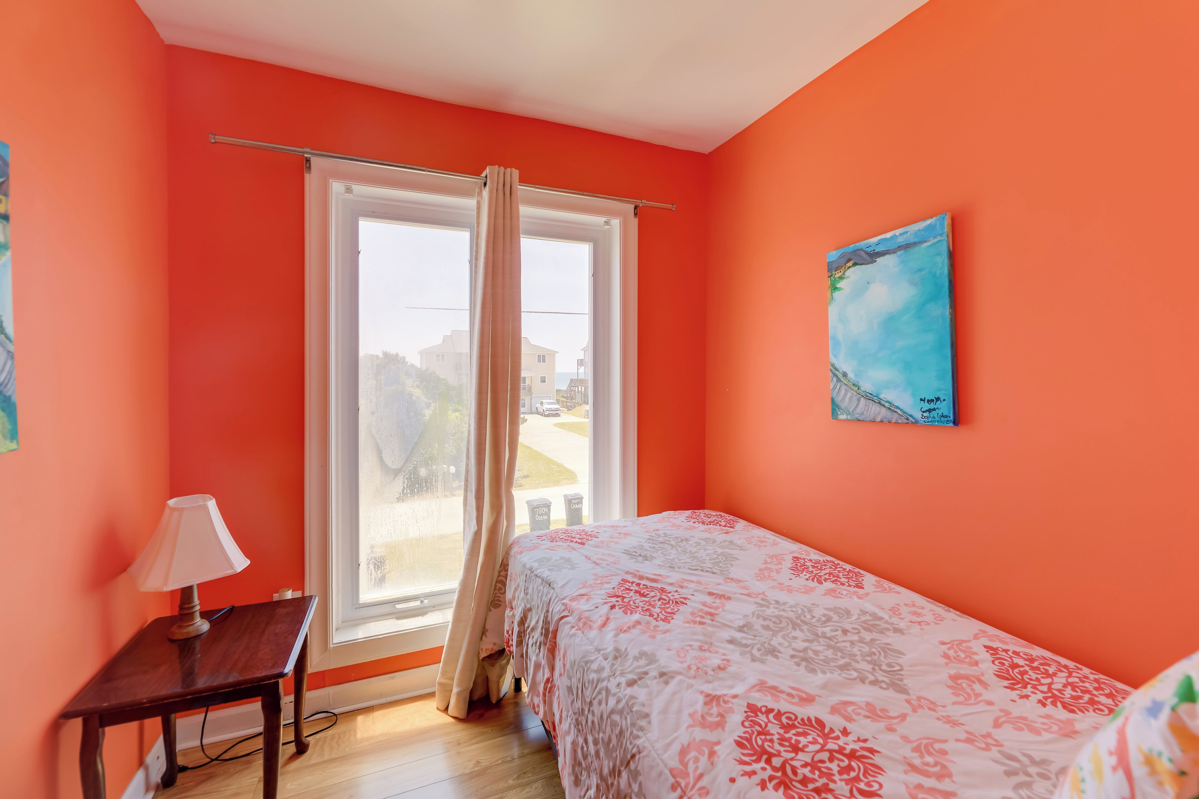 A bedroom with a bed, a window with curtains, a painting on the wall, and a small table with a lamp.