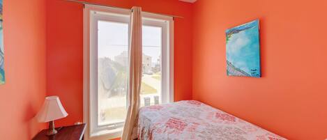 A bedroom with a bed, a window with curtains, a painting on the wall, and a small table with a lamp.
