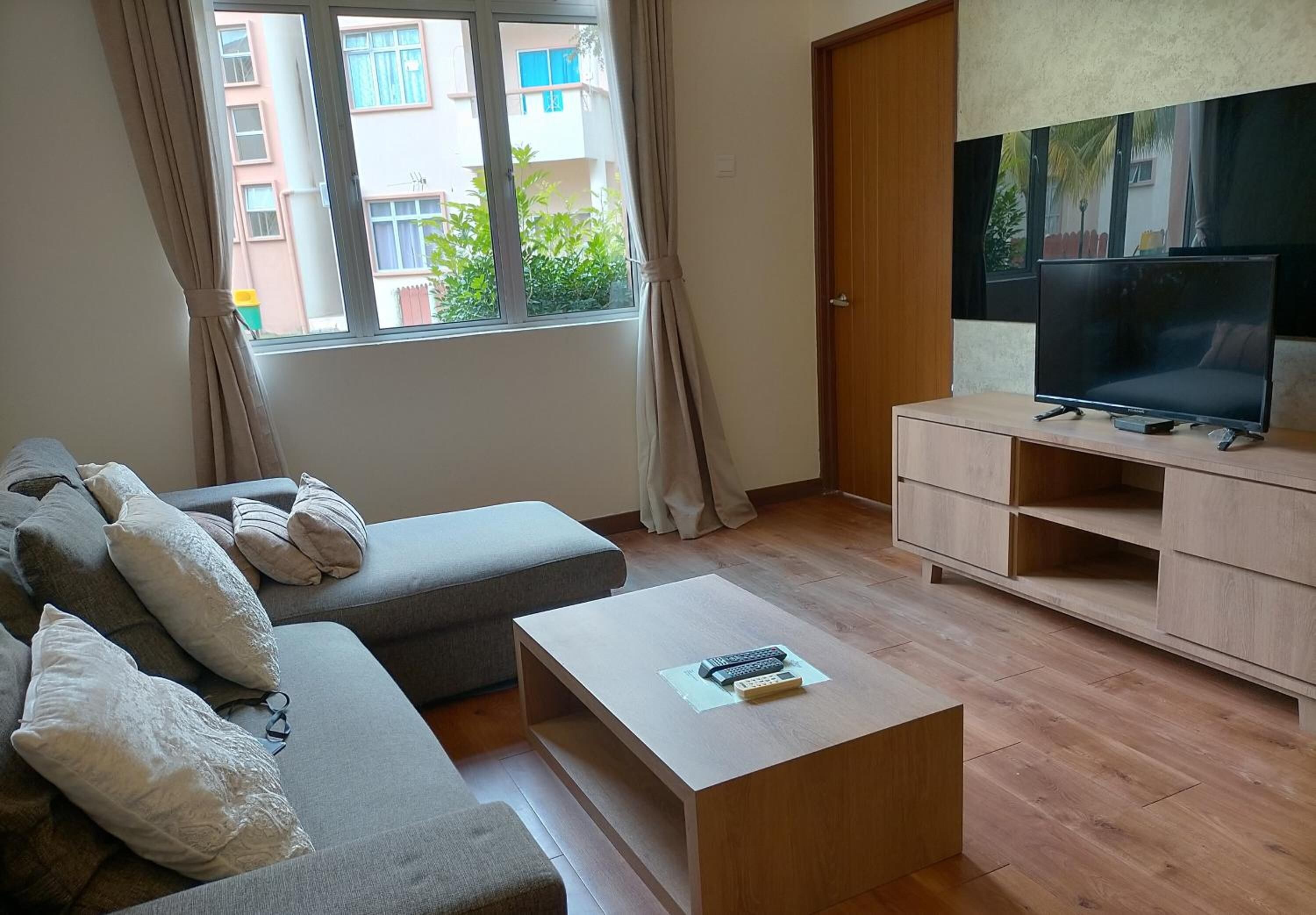 A living room with a grey sofa, a wooden coffee table, a TV on a stand, and a window with curtains.