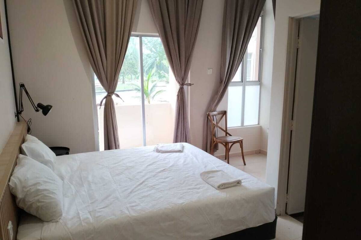 Un lit bien fait, avec du linge de lit blanc et une serviette pliée, une chaise en bois et une lampe de bureau dans une pièce dotée de grandes fenêtres et de rideaux.