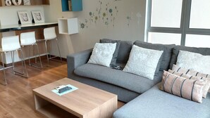 A modern living room with a grey sofa, white cushions, a wooden coffee table, and a bar area with white chairs. - Seri Bayu Resort (Sungai Pelek)
