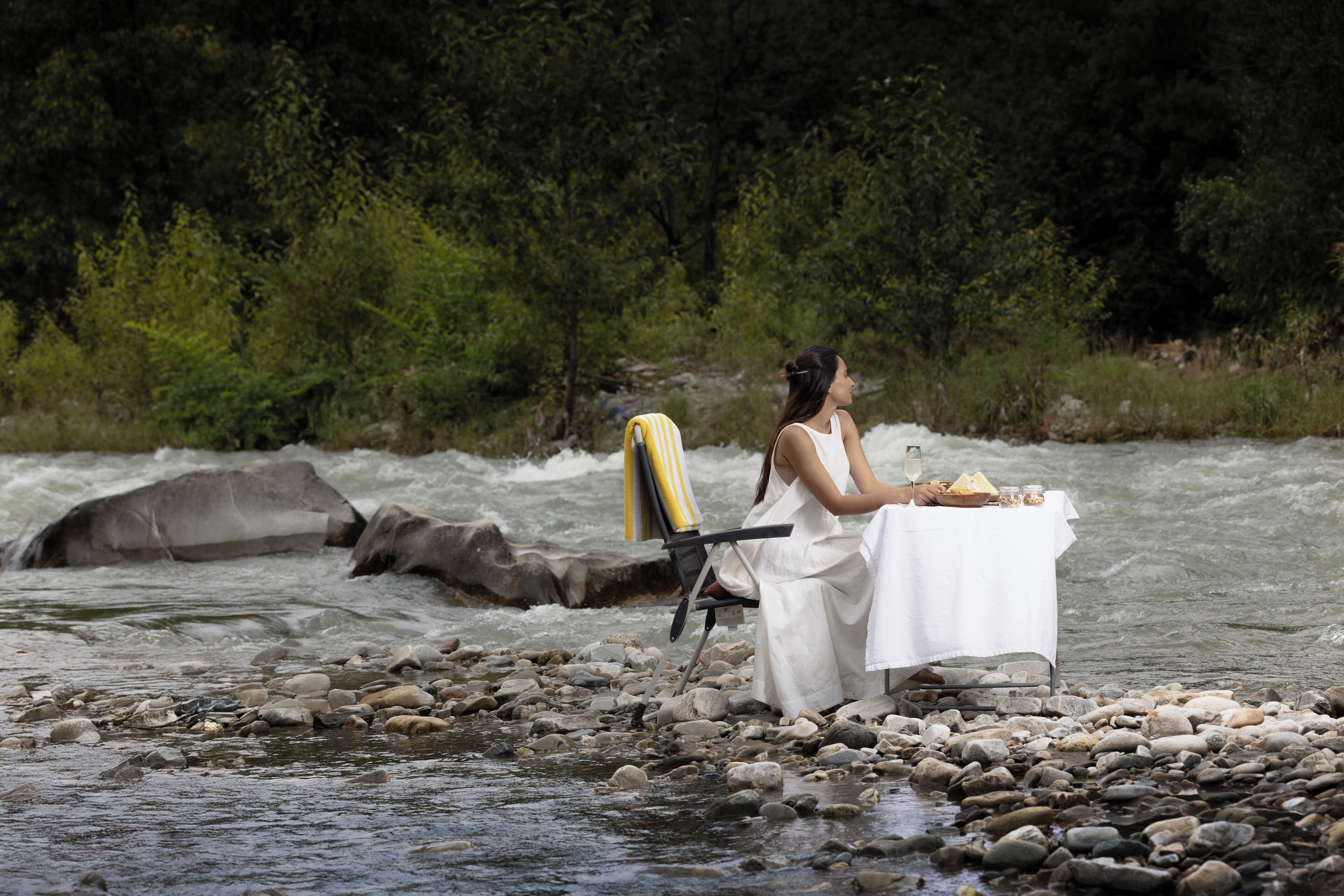 A woman sitting by a river with a table set for a meal, surrounded by rocks and greenery.