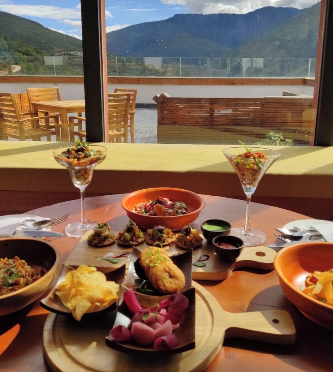 A dining table with various dishes and drinks, set against a scenic mountain view.