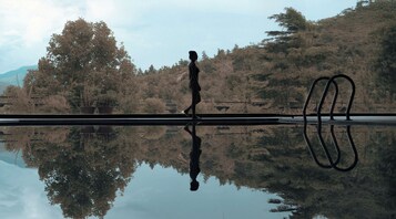 Una persona caminando junto a un cuerpo de agua tranquilo, con árboles y colinas al fondo.