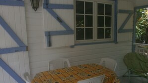 Een kamer met een raam, witte muren en blauwe accenten. Er staat een tafel met een bloemrijk tafelkleed en twee witte stoelen.