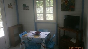 Een eetruimte met een tafel bedekt met een bloemrijk tafelkleed, blauwe stoelen, een televisie op een houten standaard en een raam met uitzicht op groen.