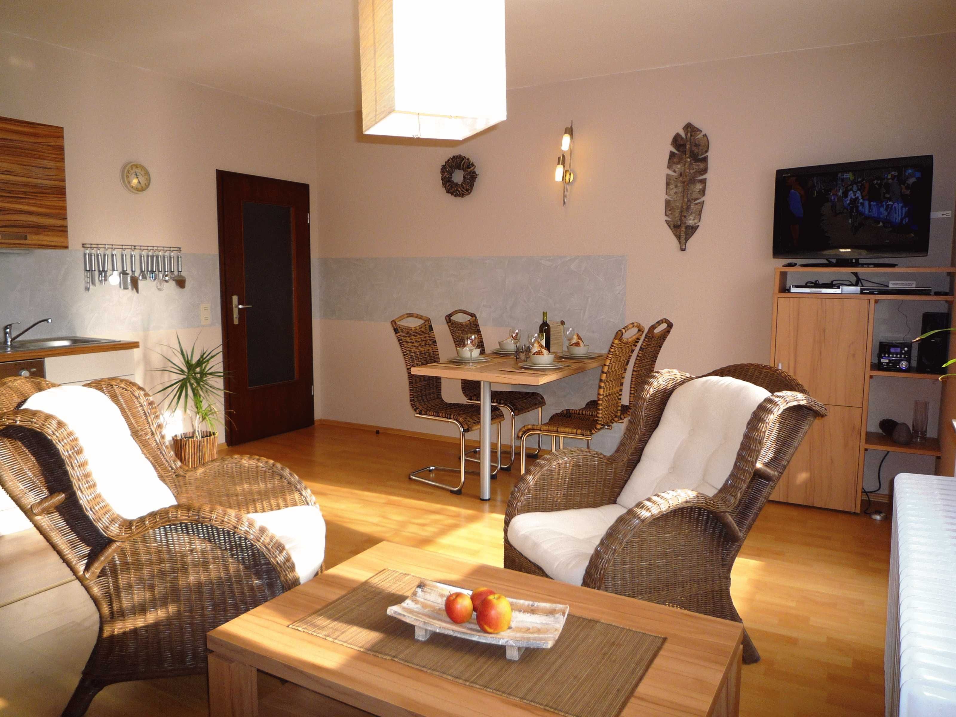 A living room with wicker furniture, a dining table set for two, and a television in the background.