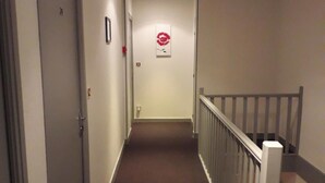 Een smalle, beklede gang met een witte deur links, een rood bord met een hart en een vogel aan de muur, en een witte leuning rechts.