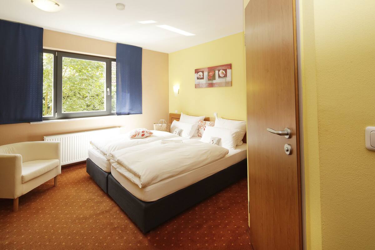 Une chambre d’hôtel comprenant un lit, une chaise, une fenêtre avec des rideaux, un radiateur et une lampe fixée au mur.