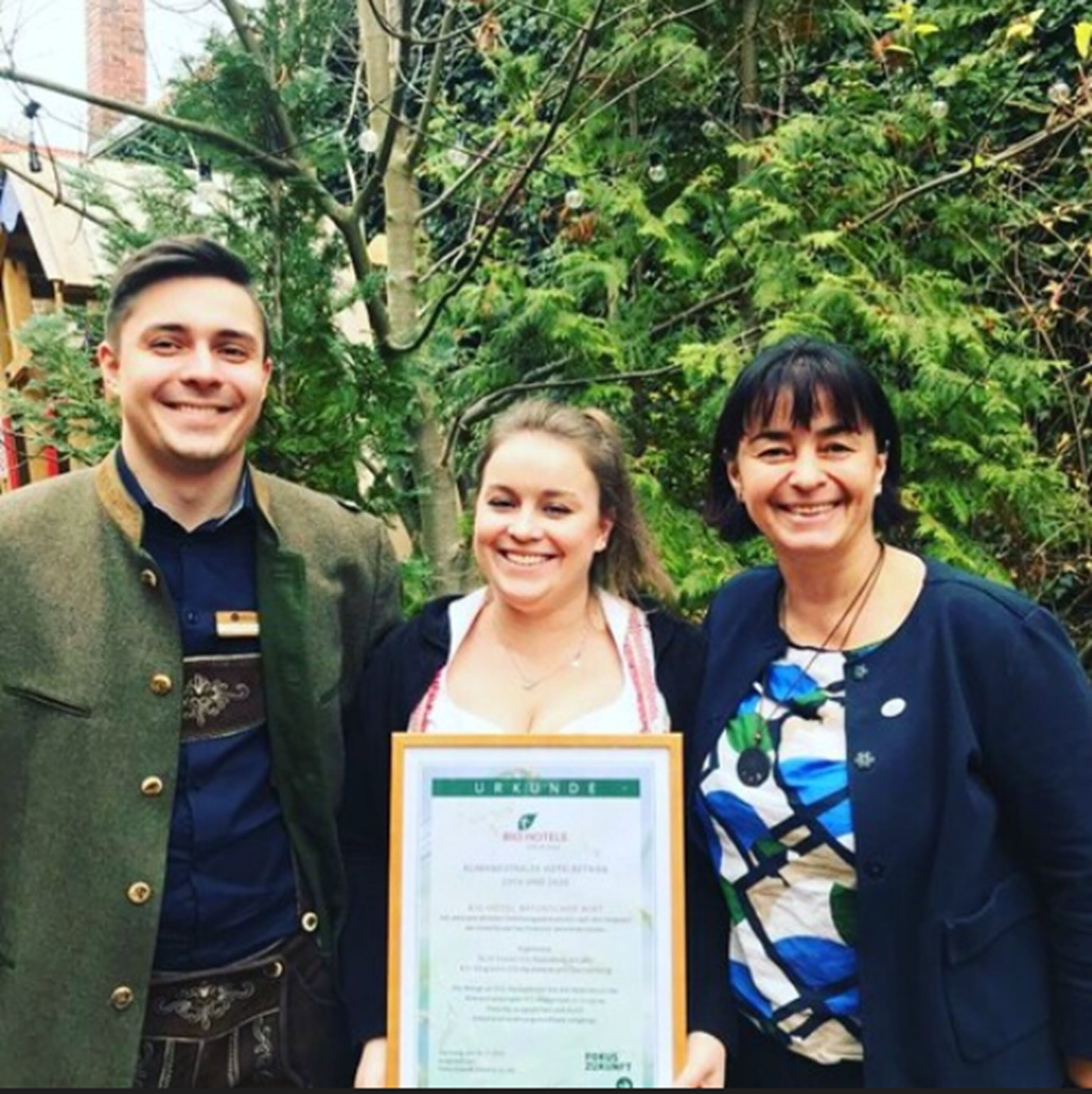 Three people holding a framed certificate outdoors with greenery in the background.