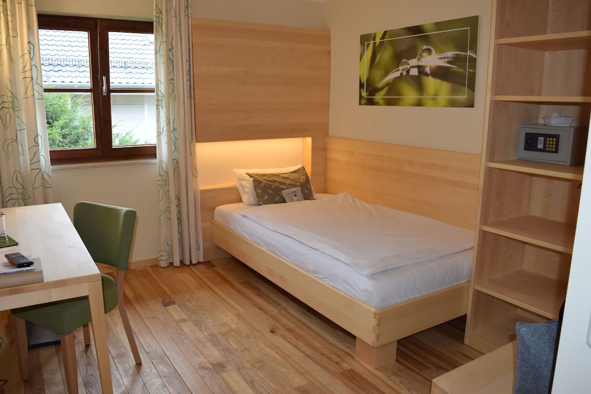 Une petite pièce agréable et chaleureuse, avec un lit simple, un bureau et une chaise. Il y a une fenêtre avec des rideaux, une étagère fixée au mur avec une radio, et un tableau au-dessus du lit.