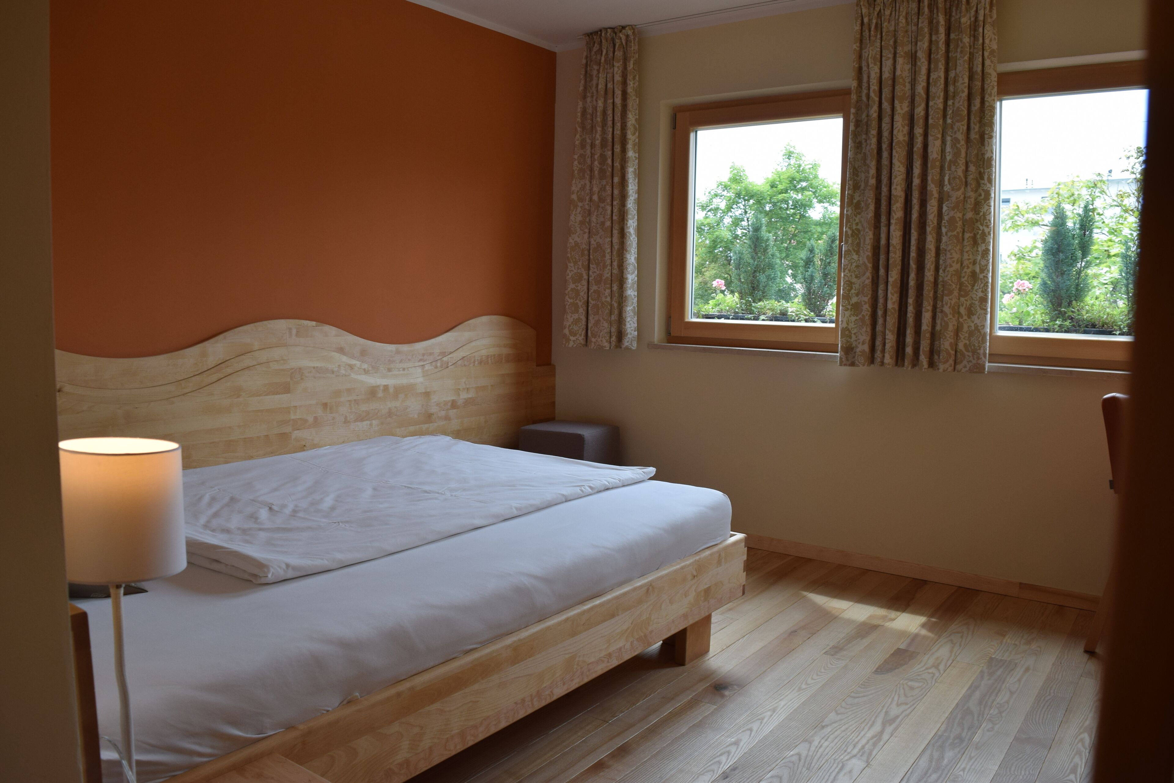 A bedroom with a wooden bed, a bedside lamp, a window with curtains, and a view of greenery outside.