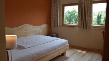 A bedroom with a wooden bed, a bedside lamp, a window with curtains, and a view of greenery outside.