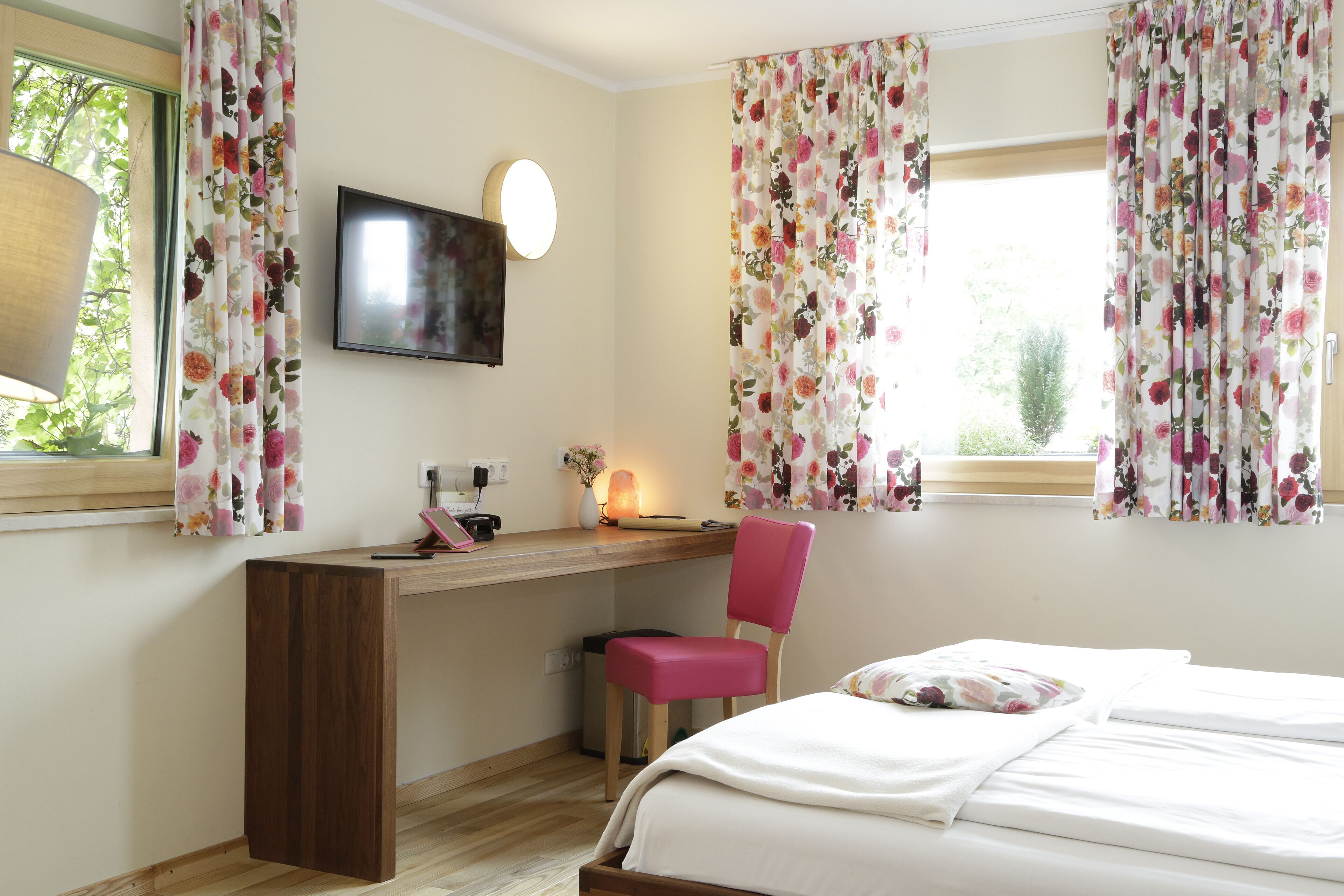 Ein Schlafzimmer mit einem Holztisch, einem rosa Stuhl, einem Bett mit weißer Bettwäsche und floralen Vorhängen.