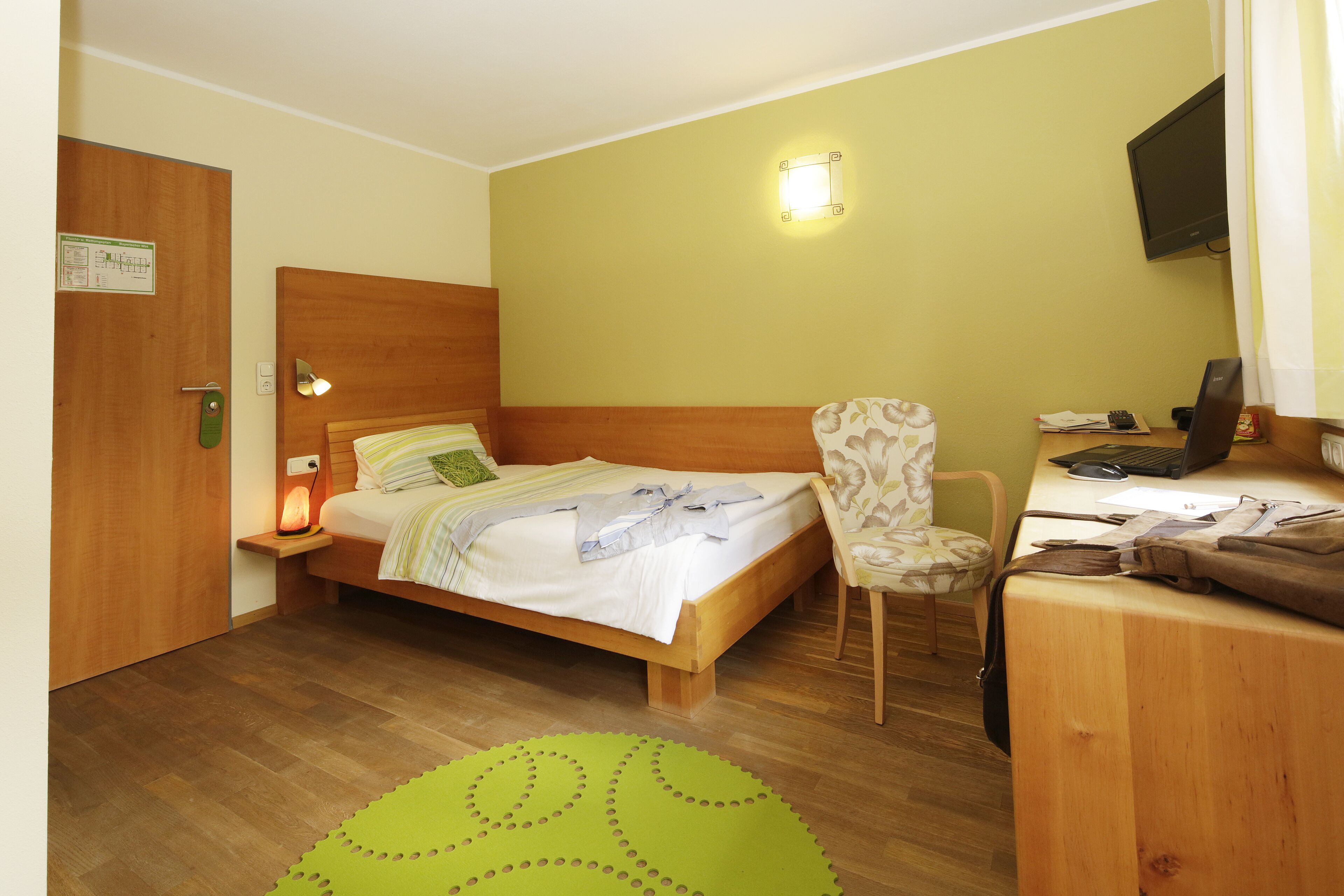 Ein Hotelzimmer mit einem hölzernen Bett, einem Schreibtisch mit Stuhl, einem grünen Teppich und einem an der Wand befestigten Fernseher.