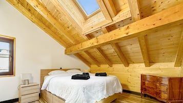 Ein Schlafzimmer mit schrägverdeckter Decke, Holzbalken, einem Bett mit weißer Bettwäsche, einem Nachttisch, einer Kommode und einem Fenster mit Blick auf den Himmel.