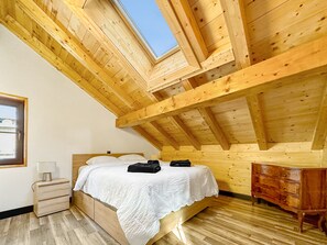Un dormitorio con techo inclinado, vigas de madera, una cama con ropa de cama blanca, una mesita de noche, un aparador y una ventana con vistas al cielo.
