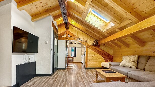 A spacious loft with a high ceiling, wooden beams, a TV mounted on the wall, a sofa, and a staircase leading to an upper level.