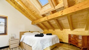 A bedroom with a sloped ceiling, wooden beams, a bed with white bedding, a nightstand, a dresser, and a window with a view of the sky.