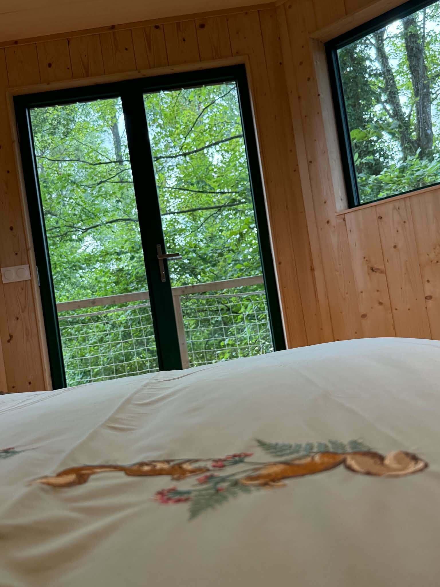 A bedroom with a large bed, wooden walls, and a view of a forest through two windows.