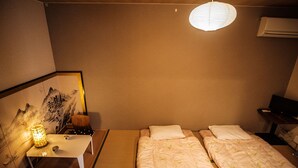 A hotel room with two beds, a desk with a lamp, a wall-mounted air conditioner, and a framed artwork.