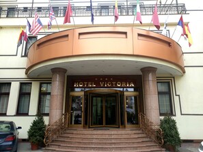La entrada del Hotel Victoria con un arco monumental, una escalera que conduce a la entrada y varias banderas exhibidas arriba.