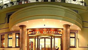 Ein Hotel Eingang mit einem Schild, auf dem 'HOTEL VICTORIA' steht, und einer Drehtür. Es gibt Stufen, die zu diesem Eingang führen, und Blumentöpfe sind in der Umgebung aufgestellt.