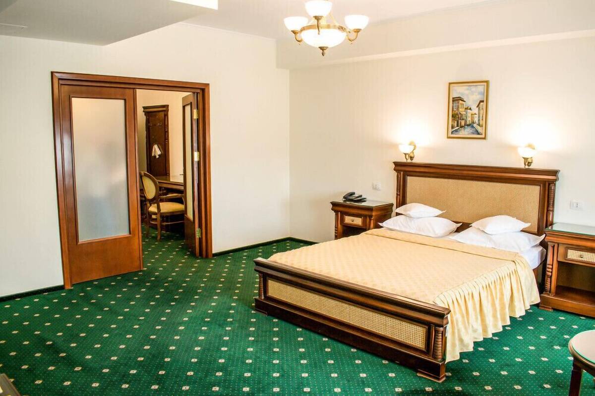 A hotel room with a large bed, a wooden headboard, a green patterned carpet, a chandelier, and a painting on the wall.