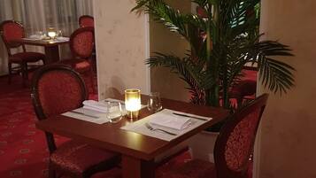 A dining area with a table set for two, red upholstered chairs, a potted plant, and a lit table lamp.