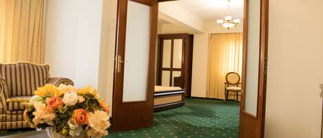 Habitación de hotel con alfombra verde, puerta de madera, cama con cabecera blanca, una silla y un florero con flores sobre una mesa.